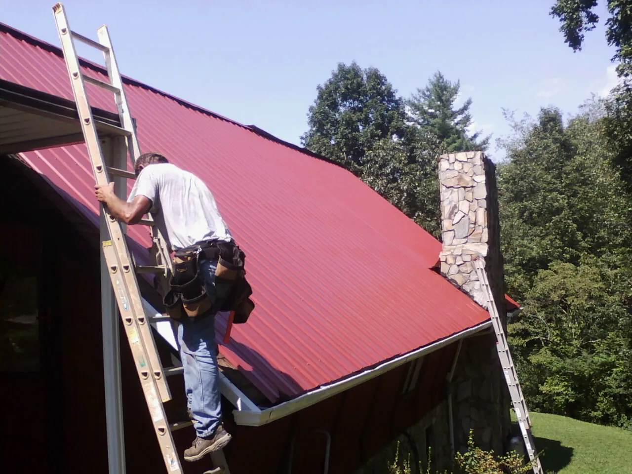 Roofer on a ladder beside a red metal roof and stone chimney in Wilmington